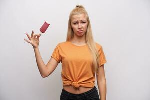 Upset young long haired blonde woman in casual clothes frowning her face and looking at camera with pout while holding ice-cream on stick, posing over white background photo