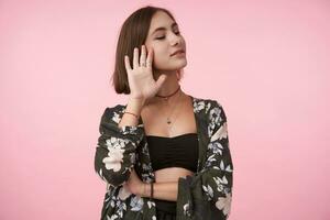 Pleasant looking young positive brown haired woman dressed in flowered shirt and black top showing raised palm and smiling slightly while posing over pink background with closed eyes photo