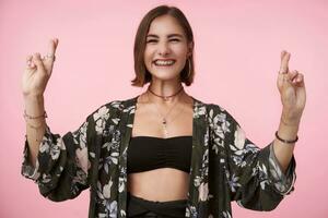 Cheerful young pretty brunette female with bob haircut smiling joyfully while raising hands with crossed fingers, standing over pink background in trendy wear photo