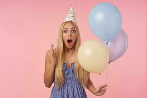 sorprendido atractivo largo peludo rubia dama en festivo ropa posando en multicolor aire globos, posando terminado rosado antecedentes con asombrado cara y redondeo ojos foto