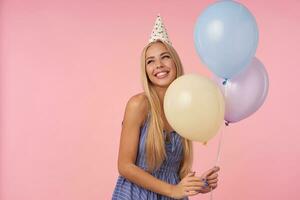 Cheerful pretty young blonde lady with long hair holding multicolored air balloons while standing over pink background in festive clothes, looking aside with wide sincere smile photo