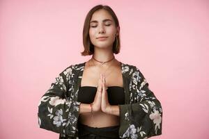 Studio shot of young attractive positive short haired lady keeping raised palms together while meditating with closed eyes, standing over pink background photo