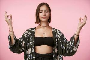 Good looking young calm short haired brunette lady keeping her eyes closed while meditating with raised hands, isolated over pink background in trendy clothes photo