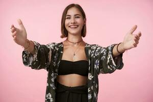 Studio shot of young pleased brown haired female with short haircut keeping her arms wide opened while smiling cheerfully at camera, isolated over pink background photo