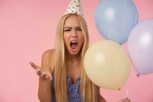 Indoor photo of severe young blonde female raising palm angrily and looking at camera with wide opened mouth, being in bad spirit, posing over pink background with bunch of helium balloons