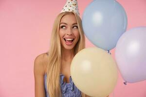 Portrait of young delighted woman with long blonde hair posing in multicolored air balloons, celebrating holiday, rejoicing nice party together with friends, standing over pink background photo