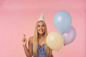 Happy attractive young blonde female making wish and crossing fingers for good luck, holding bunch of helium balloons while posing over pink background in blue summer dress and birthday cap photo