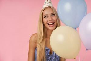 Close-up of charming young blonde female looking aside and smiling widely, posing in multicolored air balloons in blue summer dress and birthday cap, isolated over pink background photo