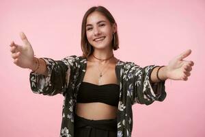 Cheerful young attractive short haired brunette woman with natural makeup smiling happily at camera while standing over pink background with opened arms photo