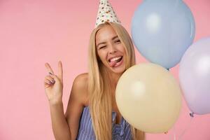 Funny shot of long haired blonde female in blue summer dress and birthday cap standing over pink background, winking at camera with sticking out tongue and raising vistory gesture photo