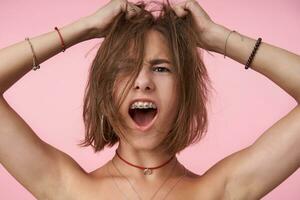 Expressive young short haired brown-eyed lady with short haircut looking excitedly at camera with opened mouth while cluthing her head with raised hands, isolated over pink background photo