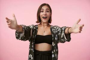 Excited young attractive brown-eyed brunette woman with short haircut looking emotionally at camera while going to give hug someone, isolated over pink background photo