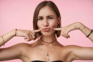 Indoor photo of young pretty brown-eyed woman with natural makeup holding index fingers on her cheeks and pursing lips while looking at camera, posing over pink background