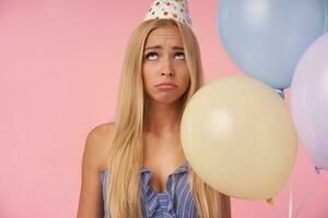 Displeased young blonde lady with long hair posing in multicolored air balloons, wearing blue summer dress and birthday cap cone hat, looking upwards and folding lips sadly photo