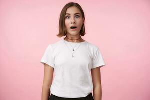 Amazed young attractive brunette female with natural makeup rounding her brown eyes while looking surprisedly at camera, standing over pink background photo