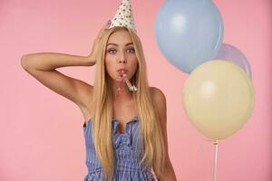 Portrait of long haired blonde lady in blue summer dress and birthday cap posing over pink background with bunch of helium balloons, raising hand to her head and looking at camera surprisedly photo