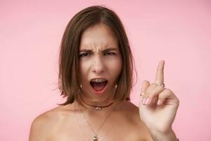 Portrait of excited young brown-eyed brunette female keeping her mouth opened and grimacing face while looking at camera, keeping forefinger raised while posing over pink background photo