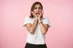 Frightened young brown-eyed brunette woman with bob haircut holding her face with raised hands while looking scaredly at camera, standing over pink background photo