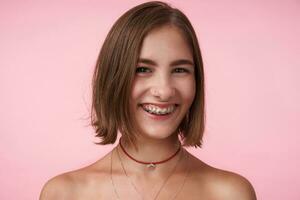 Close-up of young cheerful brown-eyed brunette female with bob haircut looking happily at camera with charming smile, isolated over pink background photo