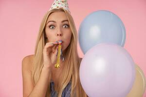 Attractive young blonde female with casual hairstyle wearing blue cone hat and blowing party horn, celebrating birthday with multicolored air balloons, having cheerful moments in her life during party photo