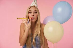 Joyful long haired blonde woman in blue summer dress and birthday cap holding bunch of helium balloons, looking at camera cheerfully and blowing party horn, isolated over pink background photo