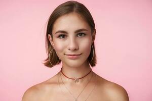 Horizontal photo of young brown-eyed brunette woman looking positively at camera and smiling slightly, standing over pink background with hands down