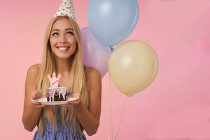 Dreamy pretty long haired blonde female keeping piece of cake with candle and smiling happily, posing over pink background with bunch of multicolored helium balloons photo