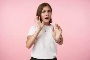 Afraid young brown haired woman with short haircut raising hands in protective gesture and looking scaredly at camera, isolated over pink background in casual wear photo