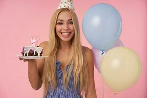 contento bonito joven rubia mujer con largo pelo havong alegre momentos en su vida durante cumpleaños fiesta, vistiendo festivo ropa y cono sombrero, en pie terminado rosado antecedentes con pedazo de pastel foto