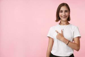 Cheerful young lovely short haired brunette female with natural makeup smiling happily while pointing aside with forefinger, isolated over pink background photo