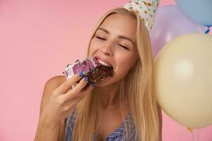 Studio shot of happy young long haired lady eating holiday cake with closed eyes, having cheerful moments in her life during birthday party, standing over pink background and helium balloons photo