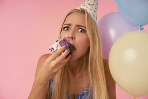 Close-up of confused young long haired blonde female in holiday cone cap eating birthday cake over multicolored air balloons, looking at camera and frowning her eyebrows photo