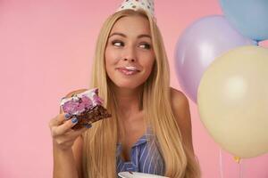 Happy attractive blonde lady smiling sincerely and looking aside with delisios cake in her hand, celebrating holiday wearing festive clothes, posing over pink background with cream on her mouth photo