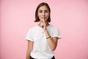 Positive young brown-eyed brunette woman with natural makeup raising hand with hush gesture and holding index finger on her mouth, isolated over pink background photo