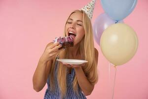 Portrait of young delighted blonde female in holiday outfit holding big piece of delicious cake and going to eat it, celebrating birthday with multicolored air balloons, posing over pink background photo