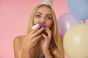 Delighted young blonde woman rejoicing while eating birthday cake, looking at camera joyfully with wide eyes opened, enjoying nice party together with friends, posing over pink background photo