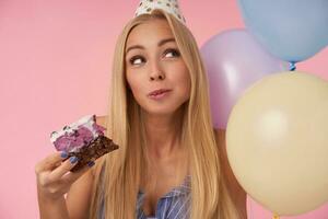 Cheerful long haired blonde woman in holiday cone hat looking aside happily while eating birthday cake, rejoicing nice party together with friends, standing over pink background photo