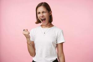 Indoor photo of expressive young brown haired woman with natural makeup keeping her mouth wide opened while screaming excitedly, isolated over pink background