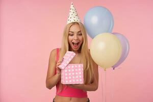 Joyful young blonde lady with long hair unpacking presents and being surprised about content, posing in multicolored air balloons over pink background, keeping her eyes and mouth wide opened photo