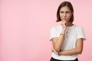 Bewildered young pretty dark haired woman dressed in basic white t-shirt twisting her mouth and frowning eyebrows while looking pensively aisde, posing over pink background photo