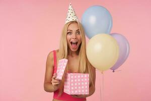 muy contento joven rubia hembra en rosado parte superior y fiesta sombrero siendo emocionado y sorprendido a obtener cumpleaños regalos, en pie terminado rosado antecedentes con manojo de helio globos foto