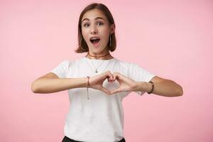 Excited young lovely dark haired woman with short haircut forming with raised hands heart and looking emotionally at camera, isolated over pink background photo
