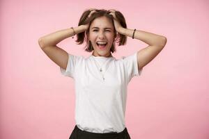 Joyful young lovely dark haired woman in casual clothes clutching her head with raised hands and looking cheerfully at camera, isolated over pink background photo