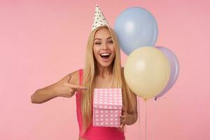 Attractive young long haired womanin pink top and cone hat celebrating holiday, holding gift-wrapped box and baing excited what is inside, isolated over pink background, looking at camera joyfully photo