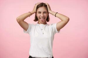 Studio photo of cheerful young pretty short haired brunette woman holding raised hands on her head and sticking happily out her tongue while posing over pink background