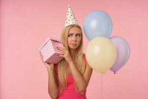 perplejo largo peludo rubia dama con casual peinado participación presente caja en manos y molesto a encontrar fuera qué es adentro, celebrando cumpleaños con multicolor aire globos, posando terminado rosado antecedentes foto