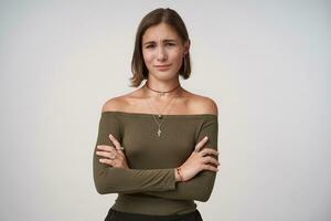 Studio photo of young attractive short haired lady with natural makeup keeping her lips folded while standing over white background with crossed hands