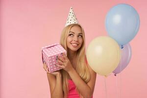 Portrait of cheerful young blonde female in pink top and birthday cap posing in multicolored air balloons, looking aside happily and keeping present box in hands, standing over pink background photo