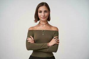 Positive young lovely short haired brunette lady smiling slightly while looking at camera and folding hands on her chest, isolated over white background photo