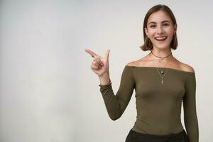 Joyful young pretty dark haired female with casual hairstyle smiling widely while showing cheerfully aside with forefinger, standing over white background photo